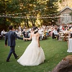 Market string lighting over guest tables at Meadowood resort. Shows bride and groom walking towards guests who are seated at outdoor tables on the lawn.