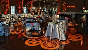 A gear pattern is projected onto the floor of an old war building called the Clock Tower. The floor is a heavily varnished oak wood, recently polished. The gear projection is in a warm tone, close to orange. The pattern is also cast over several small tables that have white linens. There is uplighting surrounding the large room, also in an orange hue. On a wall in the right upper corner of the image is an Arts Benicia logo gobo projected in a warm hue.