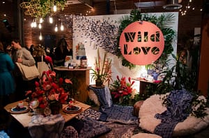 Freshbash at Beatnik Studios. Vignette shows a close up of the Wild Love circular sign. The sign has the words Wild Love knocked out to expose a backdrop of green sprigs, branches and origami birds. There's a lot of texture in the foreground with various textiles and plant arrangements. The edison pendant display hangs from the geometric fixture in the upper left hand corner.