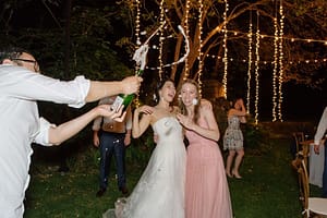 Wedding reception at Meadowood Resort with custom twinkle light backdrop hanging from large trees behind guest tables. Bride and guest are standing smiling with champagne bottle being opened with champagne shooting into the air at night.