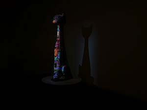 A ceramic sculpture with a cat head elongated neck and long body sits upright with a colorful pattern projected with projection mapping on the front of the cat. The cat has a soft glow of white light behind it. There are bright pinks, blues, greens and turquoise color lights shining on the cat.